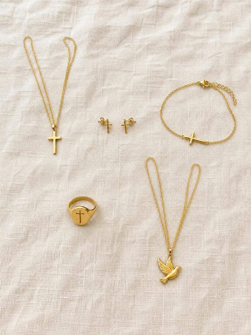 Five gold tone faith jewelry pieces on linen: cross necklace, cross studs, sideways cross bracelet, cross signet ring and dove necklace